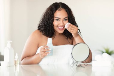 Excited plus size lady cleaning her face, using cotton pads and cleansing product, smiling at camera, bedroom interior. Body positive woman using face toner