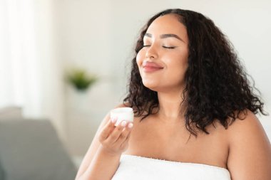 Cosmetics and skin care concept. Positive overweight lady holding cream jar and smelling, enjoying new beauty product, sitting in bedroom wrapped in towel, copy space