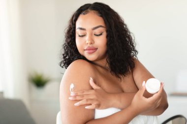 African american bodypositive lady in bath towel applying cream on shoulder, putting on moisturizing body butter at home. Black woman nourishing her skin, enjoying domestic spa procedure