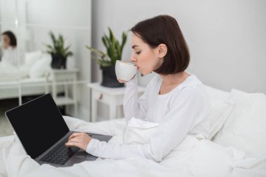 Relaxed happy beautiful brunette young woman in white pajamas chilling in bed with modern laptop, typing on computer keyboard with black empty screen, drinking coffee, mockup, copy space