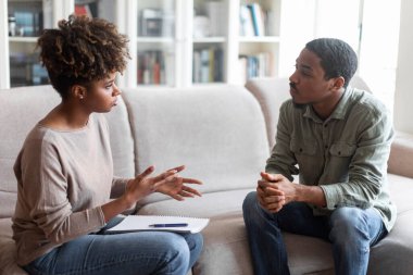 Millennial african american young woman therapist and millennial guy patient sitting on couch at counselor office, have conversation, copy space between them. Therapy, psychological help concept