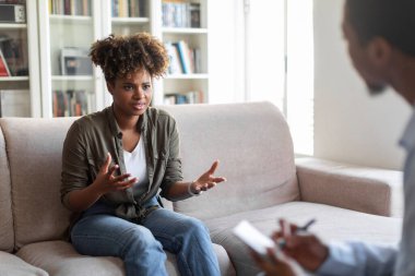 Emotional african american millennial woman in casual reclining on couch at therapist office, sharing feelings and gesturing, young black lady having therapy session with man psychologist, free space