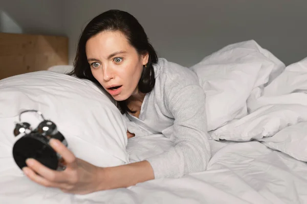 Shocked frightened young european woman lies on white comfortable bed, wake up, looks at alarm clock in bedroom interior, close up. Overslept, late for work, meeting at home and facial expressions