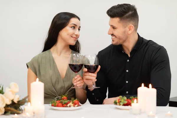 Loving hispanic millennial man and woman wearing nice festive outwear cheering with glasses of red wine, looking at each other and smiling, lovers celebrating St. Valentines Day at restaurant