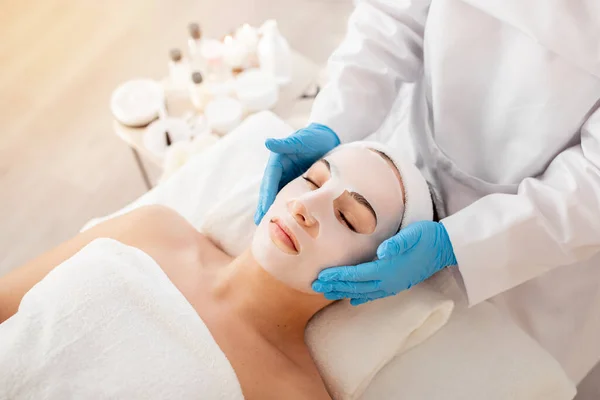 Cosmetology Concept. Beautician doctor applying face mask to young woman in spa salon, calm lady lying with closed eyes, enjoying wellness skin treatments and beauty procedures, above shot