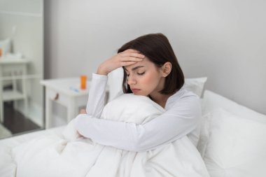 Sick young brunette woman sitting in bed under cover at home, touching head and hugging her knees, suffering from headache or fever while have cold or flue, copy space