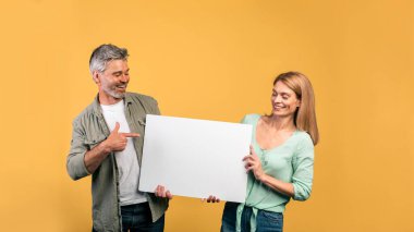 Place for your ad. Middle aged couple holding empty placard board, man pointing on it, yellow studio background. Spouses standing with white paper, free space