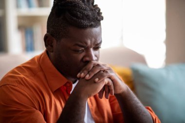Depressed millennial young black guy with stylish hairstyle in casual outfit sitting alone on couch at home, looking down, suffering from burnout, depression, closeup shot, copy space