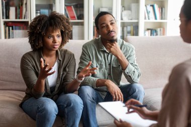 Emotional beautiful young black lady complaining about her husband at therapy session, unhappy married african american couple experiencing difficulties in relationships, visiting family counselor