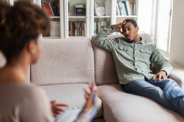 Depressed unhappy young black man reclining on couch at counselor office, listening to african american woman psychologist, millennial guy suffering from anxiety or PTSD, copy space