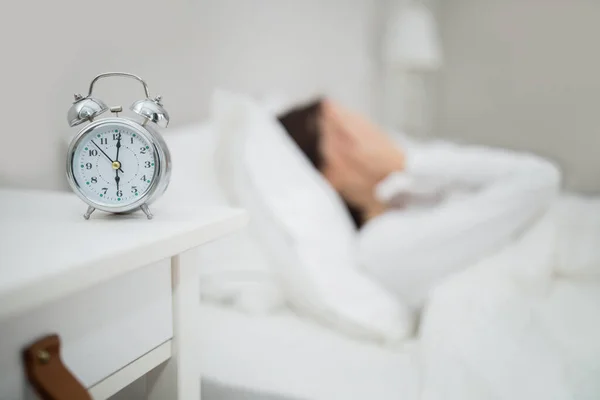 Selective focus on alarm clock over stressed young woman in white pajamas lying in bed at home, tired lady covering her face with hands, unwilling wake up early in the morning, copy space