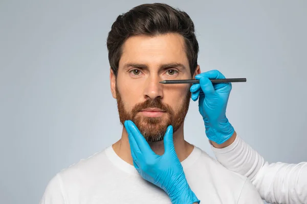 Doctor making marks on male patients face, middle aged man on consultation at surgeon, standing on grey background. Facial plastic surgery concept