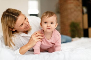 Mother playing with little baby girl while adorable toddler child crawling on bed and looking at camera. Mom enjoying child care and maternity leave