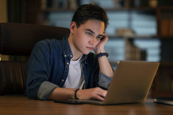 Bored unhappy young entrepreneur sitting at office desk late at night, sad guy looking at computer screen, feeling sleepy and tired, having difficulties with business, copy space. Burnout concept