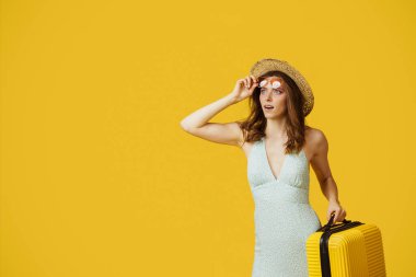 Young lady in sunglasses and summer hat holding suitcase and looking at copy space on yellow studio background. Woman getting ready for vacation