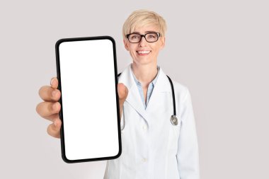 Elinde boş ekranla gülümseyen orta yaşlı kadın doktor elinde akıllı telefon, üniforma giyen arkadaş canlısı terapist bayan tıp ilanı, kolaj ve kolaj için fotokopi alanı olan boş cep telefonunu gösteriyor.
