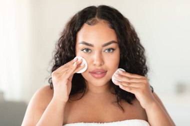 Cilt bakımı. Güzel, kilolu bir kadın, cildini temizlemek için iki pamuk pedi kullanıyor, günlük güzellik rutini yapıyor, yatak odasında havluya sarılıp oturuyor.