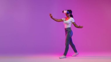 Sanal Gerçeklik Deneyimi. Amazed Black Woman VR gözlükleriyle video oyunu oynuyor, Heyecanlı Afro-Amerikan Kadın Mor Stüdyo Arkaplanı Üzerinde Yürüyor, Tam Uzunluk Çekimi, Kopyalama Boşluğu