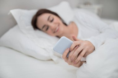 Modern akıllı telefon genç gülümseyen kadın ellerinde, beyaz pijamalı mutlu kadın sabah uyandıktan sonra yatakta uzanıyor, telefon kullanıyor, mesajları kontrol ediyor, yatak odasının içini kontrol ediyor, kopyalama alanını kopyalıyor.