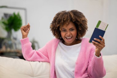 Turizm konsepti. Mutlu Afro-Amerikan bayanı rahat rahat evinde oturuyor, elini kaldırıyor ve bağırıyor, pasaport ve uçak biletlerini tutuyor, tatile çıkıyor.