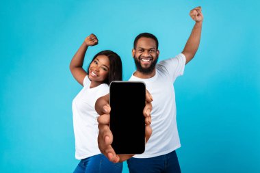Güzel uygulama. Neşeli siyah çift büyük boş akıllı telefonları gösteriyor ve başarıyı kutluyorlar. Pozitif Afrikalı Amerikalı erkek ve kadın reklam tasarımı için fotokopi alanı olan boş cep telefonlarını gösteriyorlar.