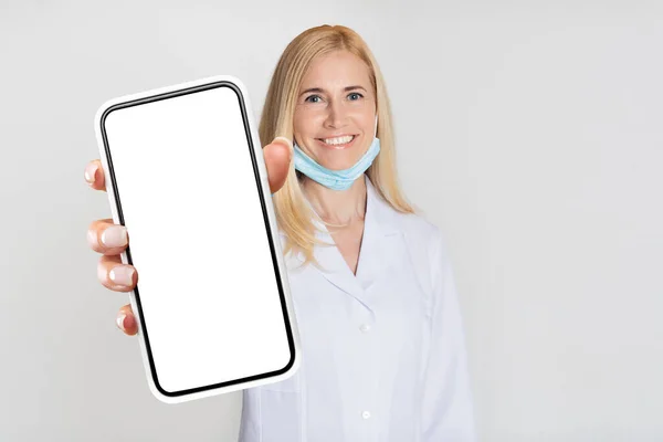 穿着制服微笑的女医生在镜头前展示带有空白白屏的手机 中年医生推荐移动应用程序进行在线医疗咨询 — 图库照片