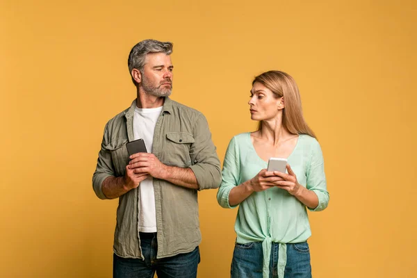 Middle aged woman spying on her husband using smartphone, texting sms or scrolling social media news feed, standing over yellow background, studio shot