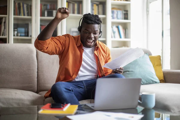 Emotional chubby young black man in casual outfit entrepreneur celebrating success, sitting on sofa, using laptop and headset, holding papers and raising hand up, working from home, copy space