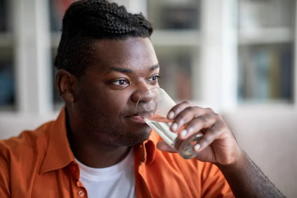Closeup shot of happy plump millennial black guy with dreadlocks in casual outfit sitting on sofa in living room, drinking water at home, looking at copy space for advertisement