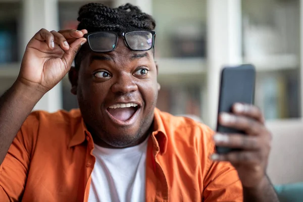 感情的な若いぽっちゃり黒人男性は興奮し 携帯電話で良いメッセージを受信し アフリカ系アメリカ人の男は ビジネスで成功を収めている携帯電話の画面を見て 眼鏡を削除し ニュースを読んで クローズアップ — ストック写真
