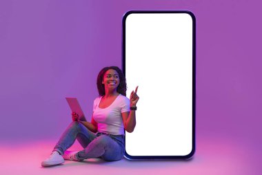 Dijital tabletli gülümseyen siyah kadın mor stüdyo arkaplanının üzerinde neon ışıkta otururken büyük boş bir akıllı telefon ekranını işaret ediyor, gülümseyen bayan yeni bir uygulama veya online teklif öneriyor,