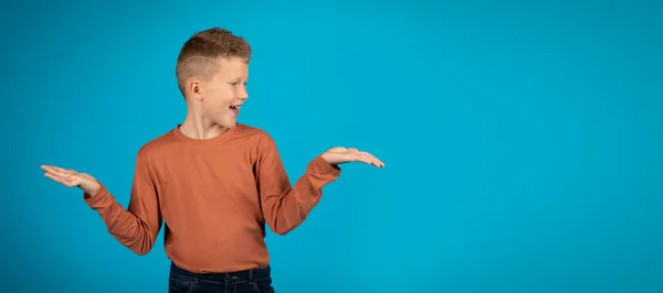 Make Choice. Cheerful preteen boy making scales with his empty palms, smiling happy male kid comparing options while standing isolated over blue studio background, panorama with copy space
