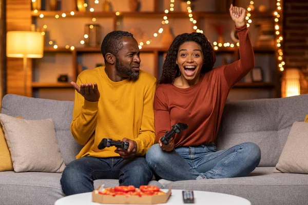 Excited cheerful pretty young black woman holding joystick raising hand up and screaming, celebrating her victory, beat her husband in video games competition, cozy home interior