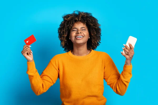 Emotional pretty young african american woman holding modern cell phone and credit card isolated on blue studio background, got bank loan online, received salary, checking bank account