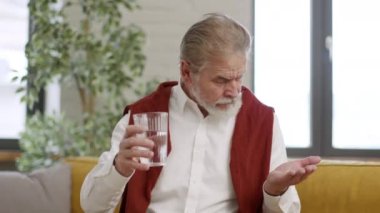 Evde tedavi ol. Sakallı sakallı sakin bir adam. İlaç alıyor ve camdan su içiyor. Kanepede dinleniyor.