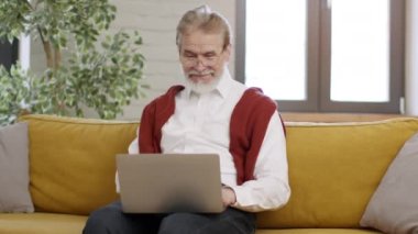 Emeklilik ve serbest çalışma. Gözlüklü, gülümseyen, uzaktan kumandalı, dizüstü bilgisayarda çalışan, kanepede oturan ve rapor okuyan, boş alan olan yaşlı adam.
