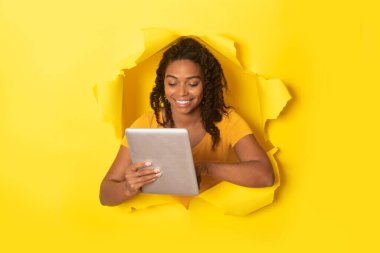 Positive black lady using digital tablet, posing in hole in torn paper on yellow background, studio shot, collage. Communication, great application concept