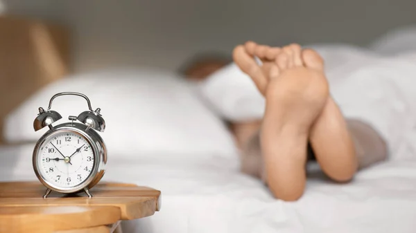 Legs of young european woman lies and sleeps on white comfortable bed, selective focus on alarm clock on table in bedroom interior, panorama. Time to wake up, overslept, late for work, meeting at home