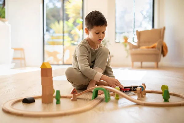 Busy serious caucasian little kid builds city, plays wooden road, train and cars in nursery room interior. Fun alone in kindergarten, entertainment at home, fantasy and childhood, education with game