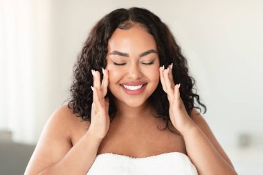 Happy oversize woman touching her face and smiling with closed eyes, making beauty treatments at home. Young body positive lady enjoying selfcare