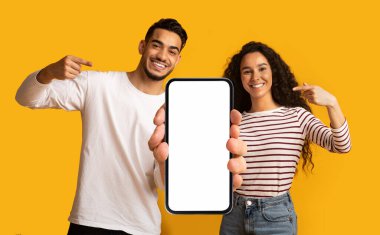 Place For Ad. Cheerful Middle Eastern Couple Demonstrating Big Blank Smartphone With White Screen For Mockup And Pointing At It, Happy Arab Man And Woman Advertising New Mobile App Or Website