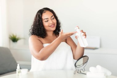 Happy black body positive lady applying moisturising lotion on hand, holding bottle, woman standing wrapped in towel after bath, bedroom interior, free space