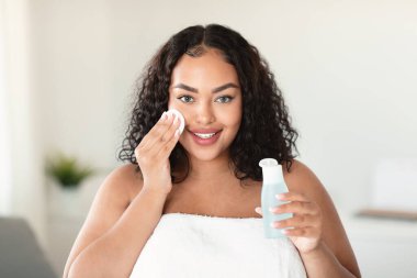 Beautiful black body positive lady cleaning her face from makeup with cotton pads and micellar water, looking and smiling at camera, standing wrapped in towel at home interior