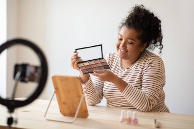 Cheerful pretty curly young chubby hispanic lady beauty blogger sitting at desk full of various makeup products, recording video on smartphone, showing eyeshadow palette, recommending cosmetics