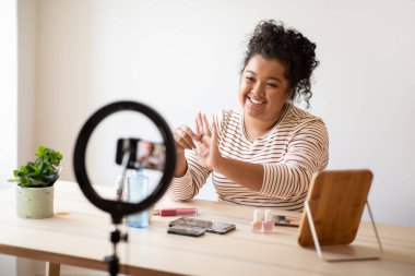 Positive friendly hispanic young lady plus size in casual outfit streaming from home, using pro blogger set and smartphone, showing brand new nail polisher, promoting cosmetics, copy space