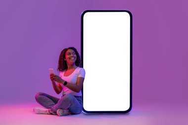Smiling black woman using mobile phone while sitting near huge blank smartphone with empty white screen, african american female posing in neon light over purple studio background, mockup