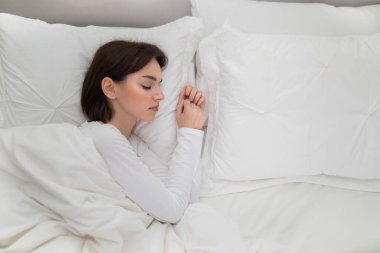 Top view of young brunette woman in white pajamas sleeping alone in big bed at home, female going through breakout, divorce, lifetime crisis, copy space for advertisement next to lady