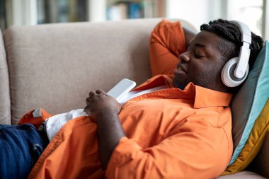 Side view of relaxed happy young overweight black man in comfy outfit chilling on couch, using wireless headphones and smartphone, listening to favorrite music at home, copy space