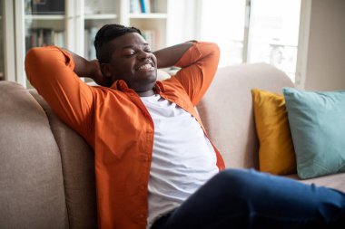 Relaxed plus size young black guy with braids in casual comfy outfit chilling alone on couch at home, holding hands behind his head and smiling with closed eyes, copy space, enjoying his new house