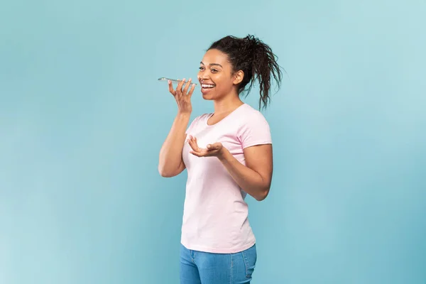 Happy young black lady using voice assistant on smartphone and gesturing, recording message while standing over blue background, studio shot, copy space